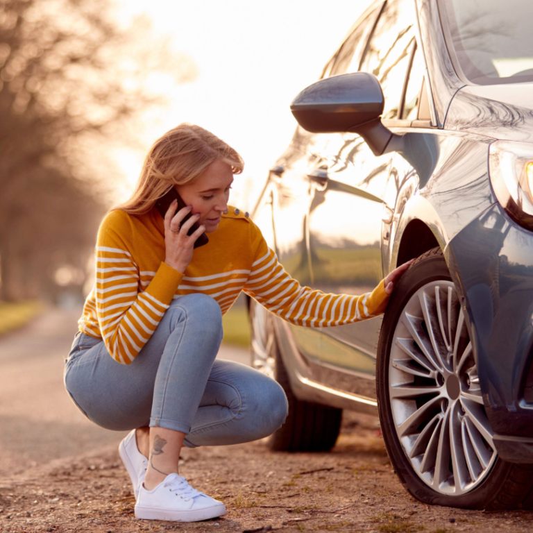 A woman in a striped, orange jumper kneels on the phone next to her car, which is stopped on the side of a country road.
